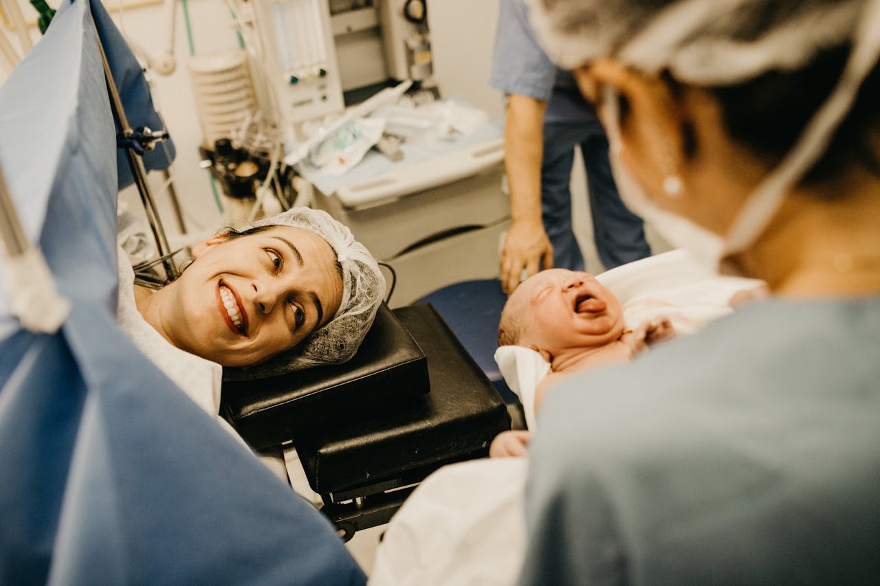 A mother smiles lovingly at her newborn in a hospital delivery room, attended by medical staff.